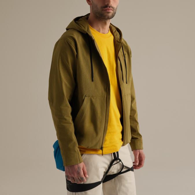 Man wearing an olive green hooded jacket, yellow t-shirt, light pants, and a climbing harness with a blue chalk bag.
