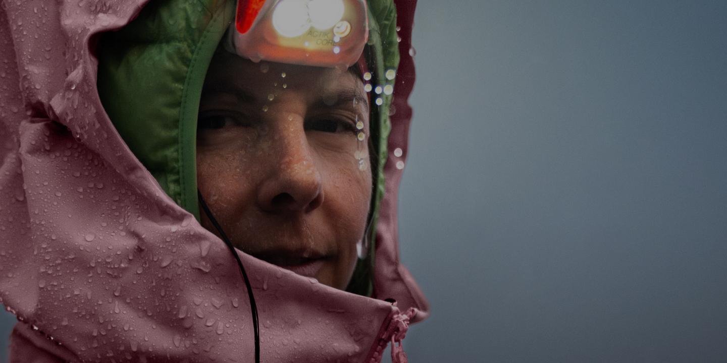 A person in a pink rain jacket with a lit headlamp, covered in water droplets.