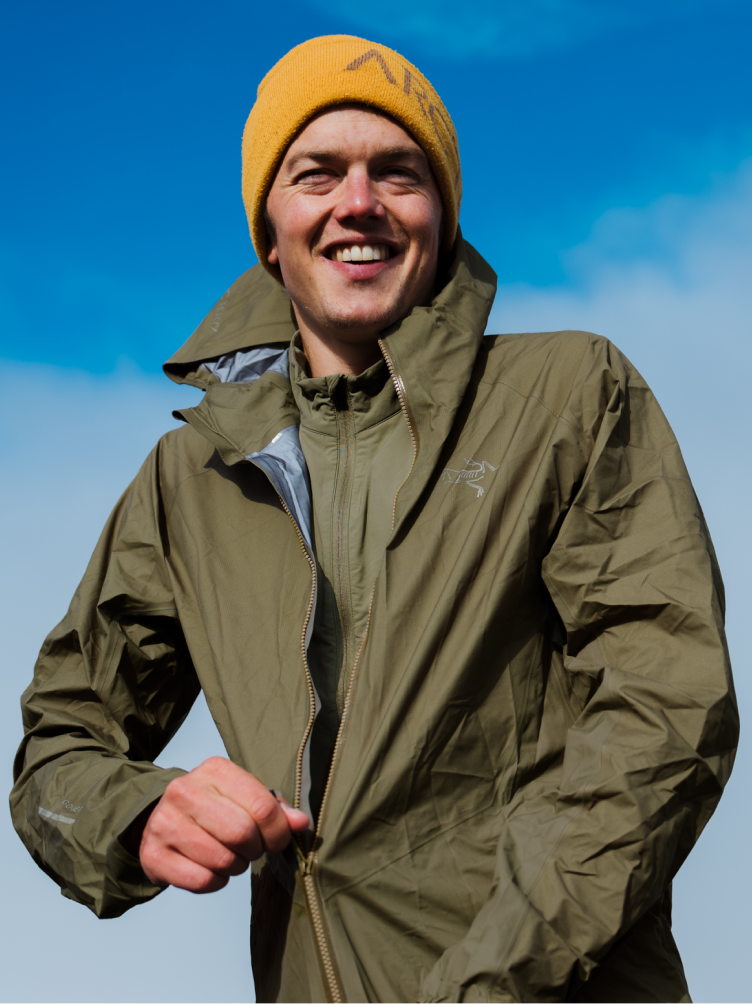 A smiling man in a yellow beanie and olive green jacket zips up his coat under a blue sky.