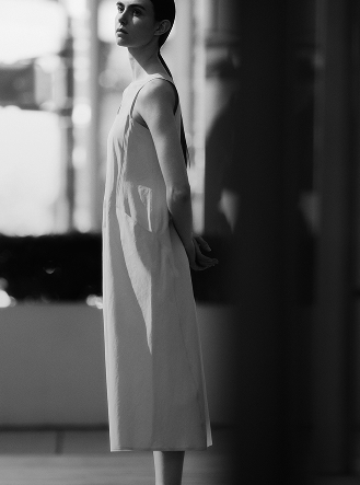 Black and white photo of a woman in a sleeveless dress, looking up with hands behind her back.