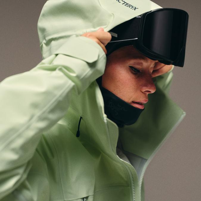 Woman in a light green Arc'teryx jacket and goggles adjusts her hood.