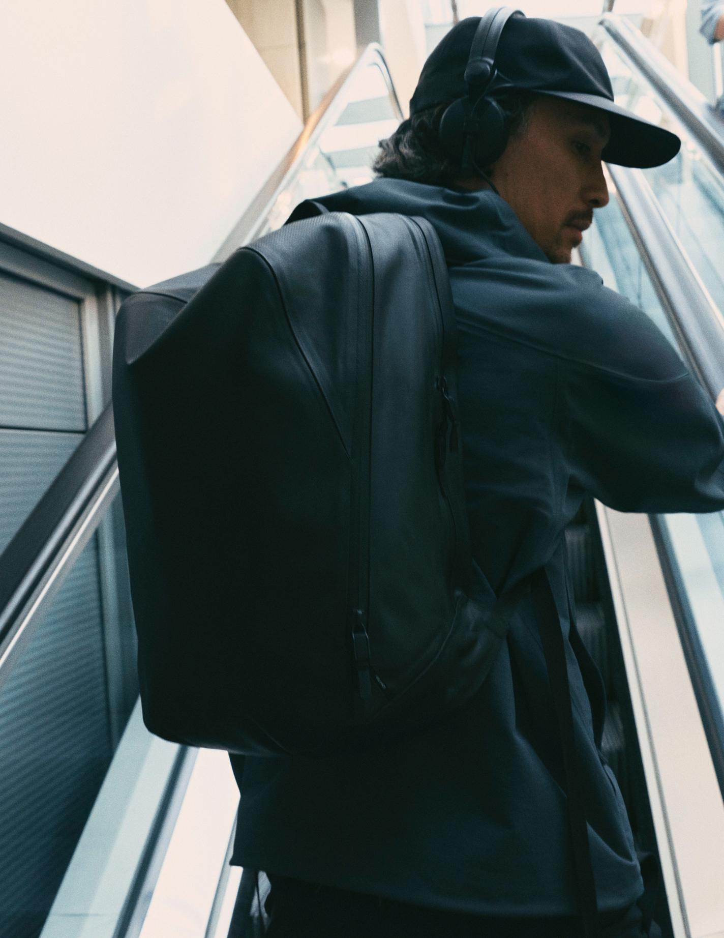 Man in a black cap and headphones, wearing a black backpack, on an escalator.
