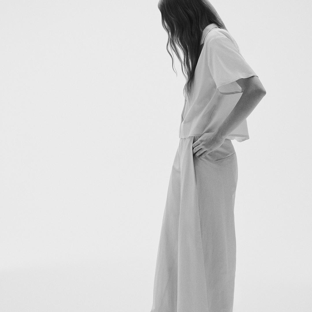 Monochromatic image of a person with head bowed, wearing a short-sleeved shirt and wide-leg pants, with one hand in a pocket.