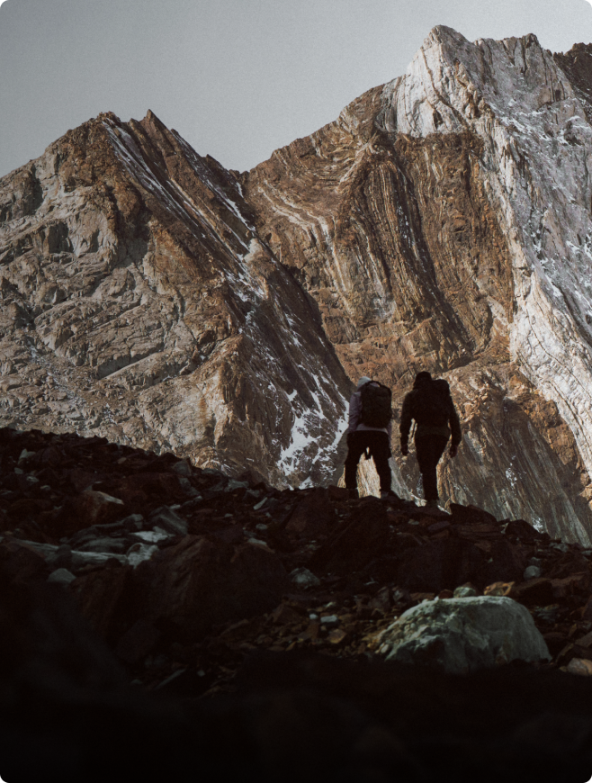 Two hikers on a rocky path look up at a vast, layered mountain with patches of snow.