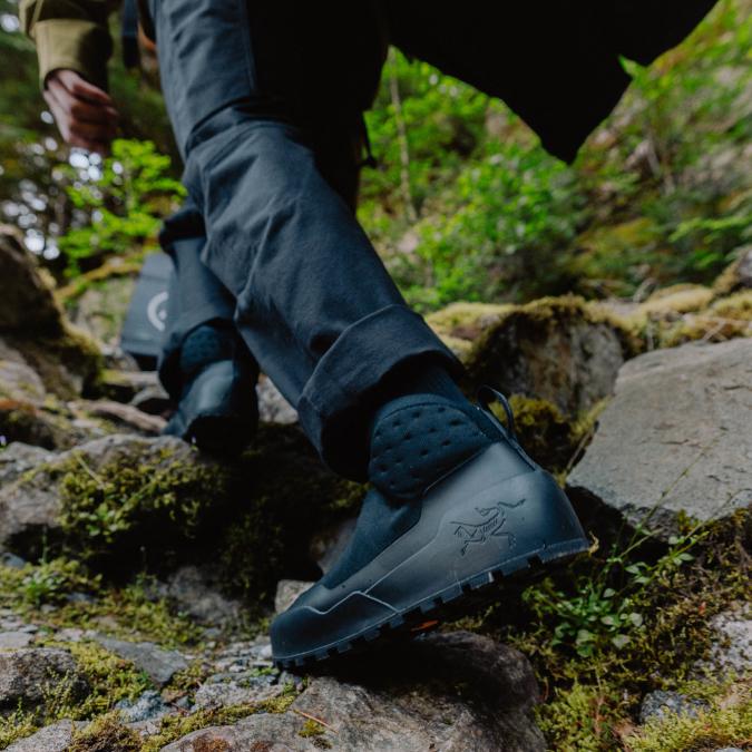 Close-up of a person wearing black Arc'teryx hiking shoes and pants, walking on a rocky, mossy trail.