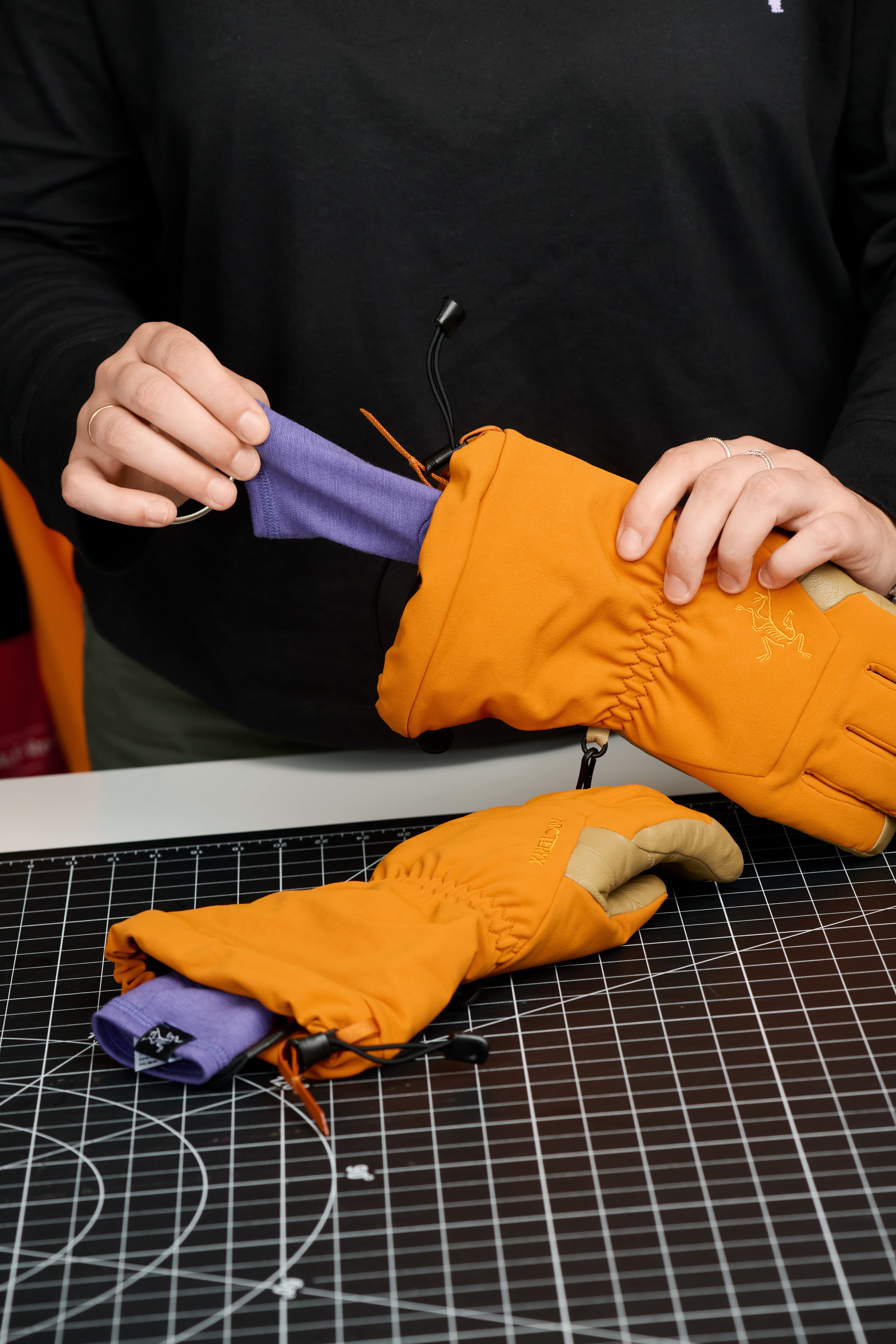 Hands putting a purple liner into an orange Arc'teryx glove, with another lined glove on a black grid mat.