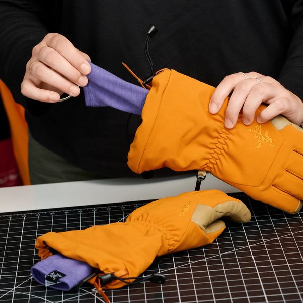 Hands putting a purple liner into an orange Arc'teryx glove, with another lined glove on a black grid mat.