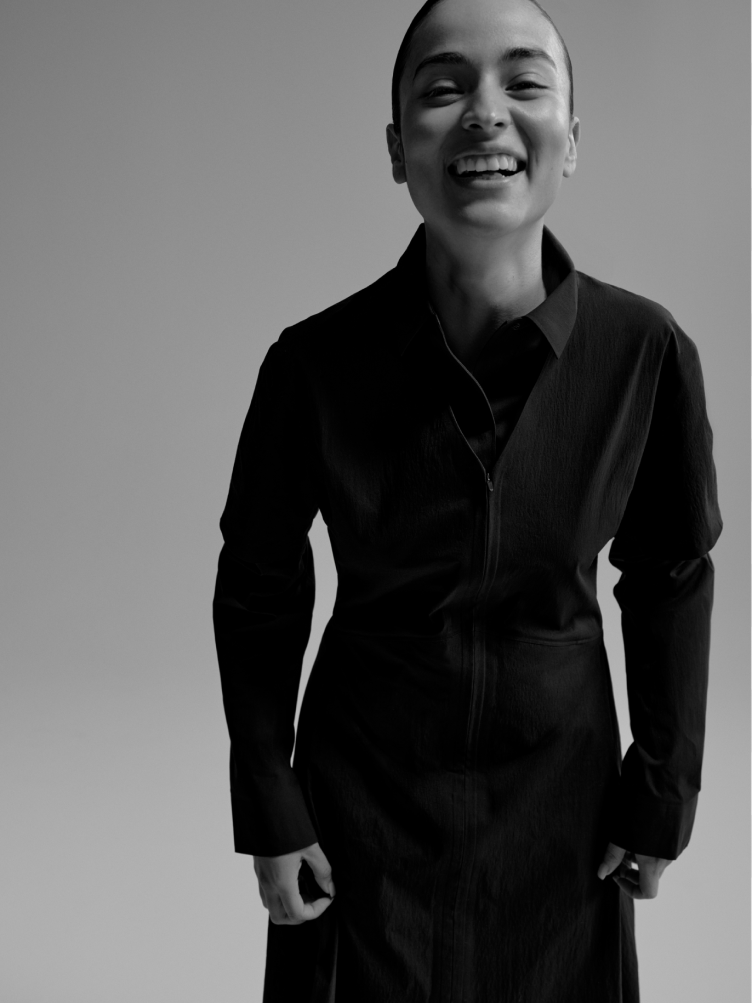 A broadly smiling person with a shaved head wears a dark zippered top in a black and white photo.