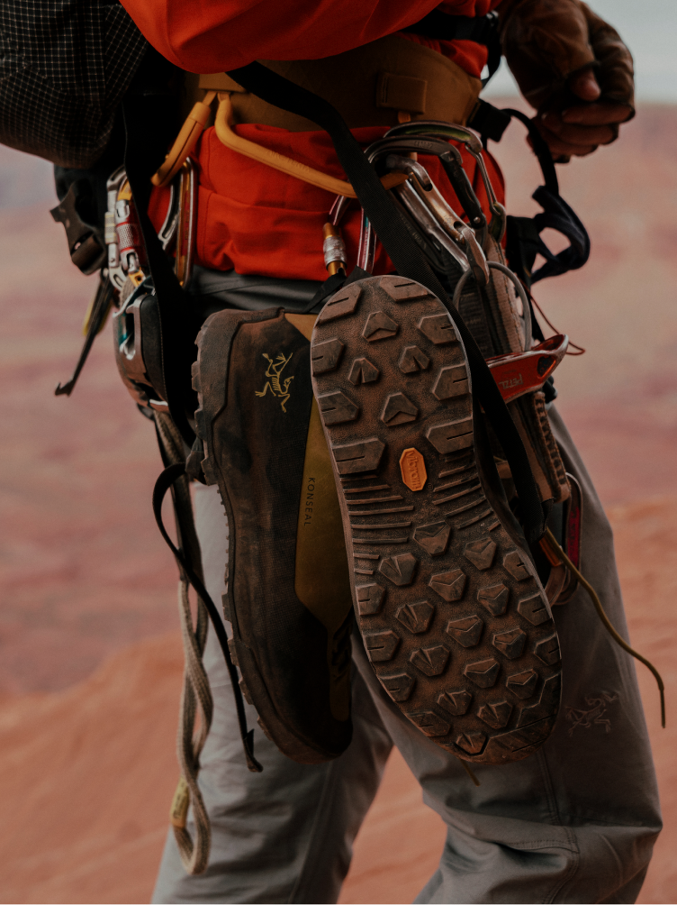 A person in climbing gear has two shoes hanging from their harness, one revealing a textured Vibram sole and the other an Arc'teryx logo.