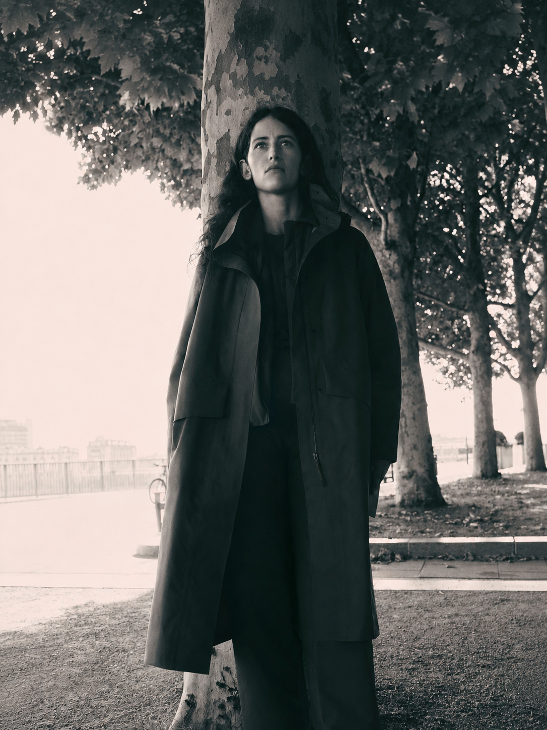 Monochrome image of a woman in a long dark coat leaning against a tree, looking upward in a park.