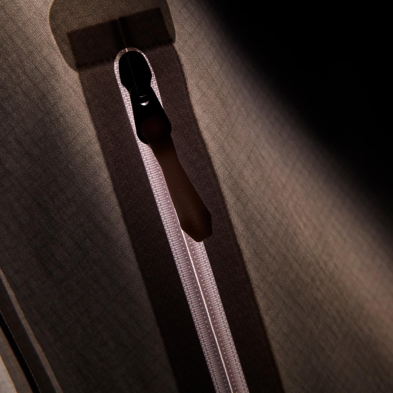 A black zipper pull and reflective zipper track against dark textured fabric, illuminated by a diagonal beam of light.