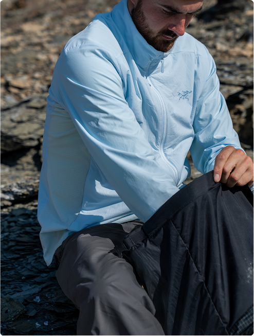 A man in a light blue Arc'teryx jacket and dark pants sits on rocky ground.