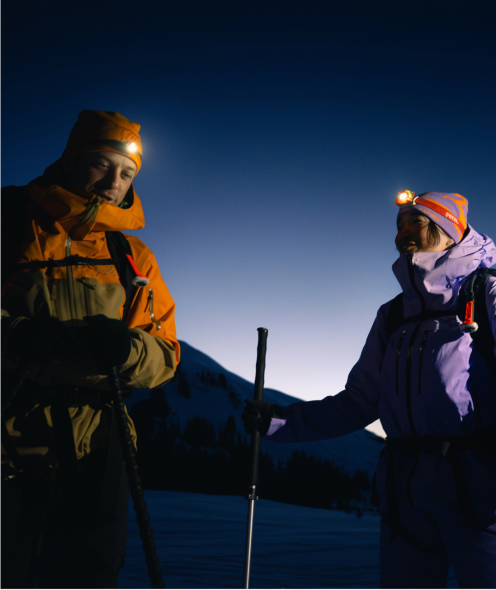 Two people in winter gear with headlamps on, standing on a snowy mountain at twilight.