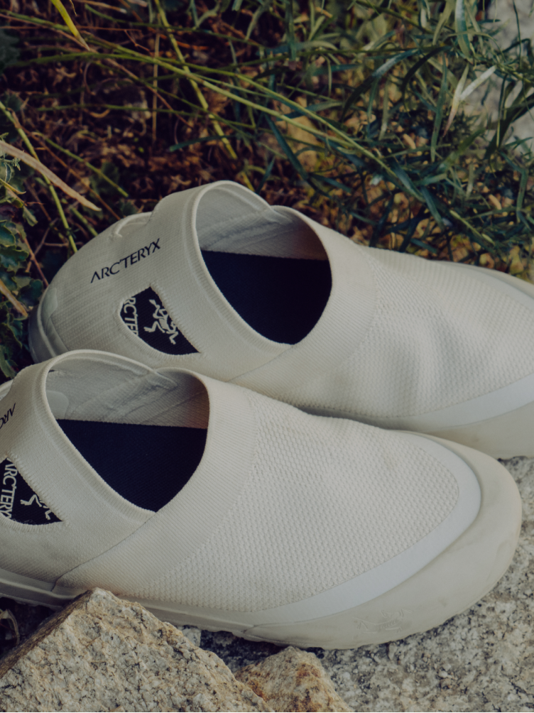 A pair of light-colored Arc'teryx slip-on shoes on rocks with green foliage.
