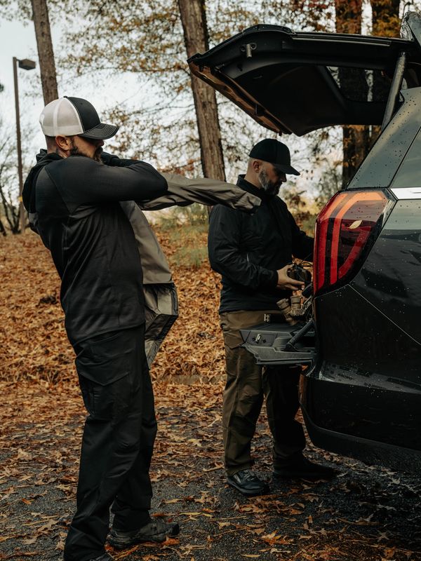 Two men, one putting on a jacket and the other by an open car trunk, stand outdoors surrounded by fallen autumn leaves.
