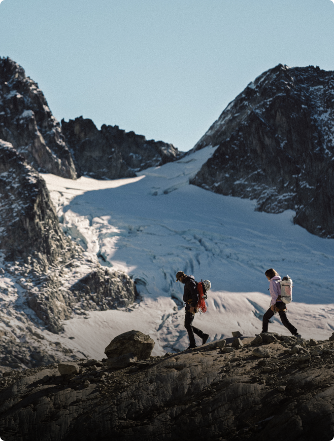 Two hikers with backpacks walk on a rocky ridge with a glacier and snowy mountains in the background.