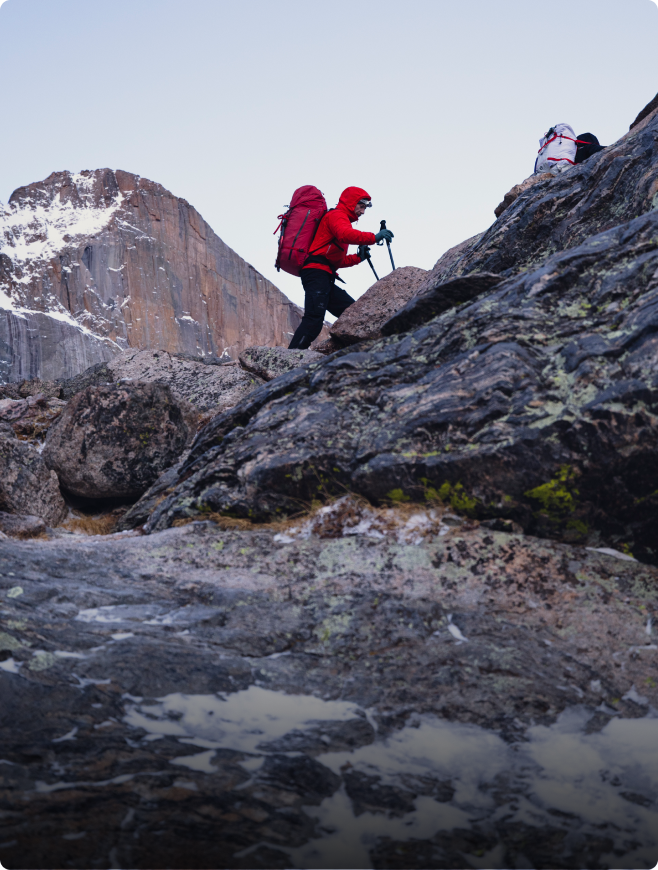 A person in a red jacket and backpack uses trekking poles to climb a rocky mountain, with a snow-capped peak in the distance.
