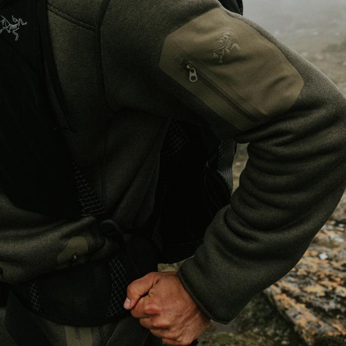 A person wearing a dark green Arc'teryx long-sleeved top with a zippered arm pocket and a backpack.
