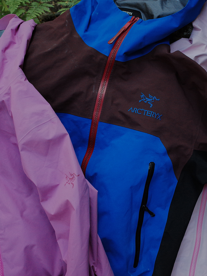 Two Arc'teryx jackets, one blue, brown, and black, and one pink, with logos visible, on a leafy surface.