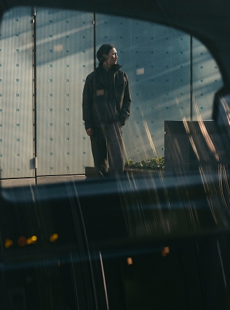 A person in dark clothing stands outside a glass building, viewed through a reflective car window.