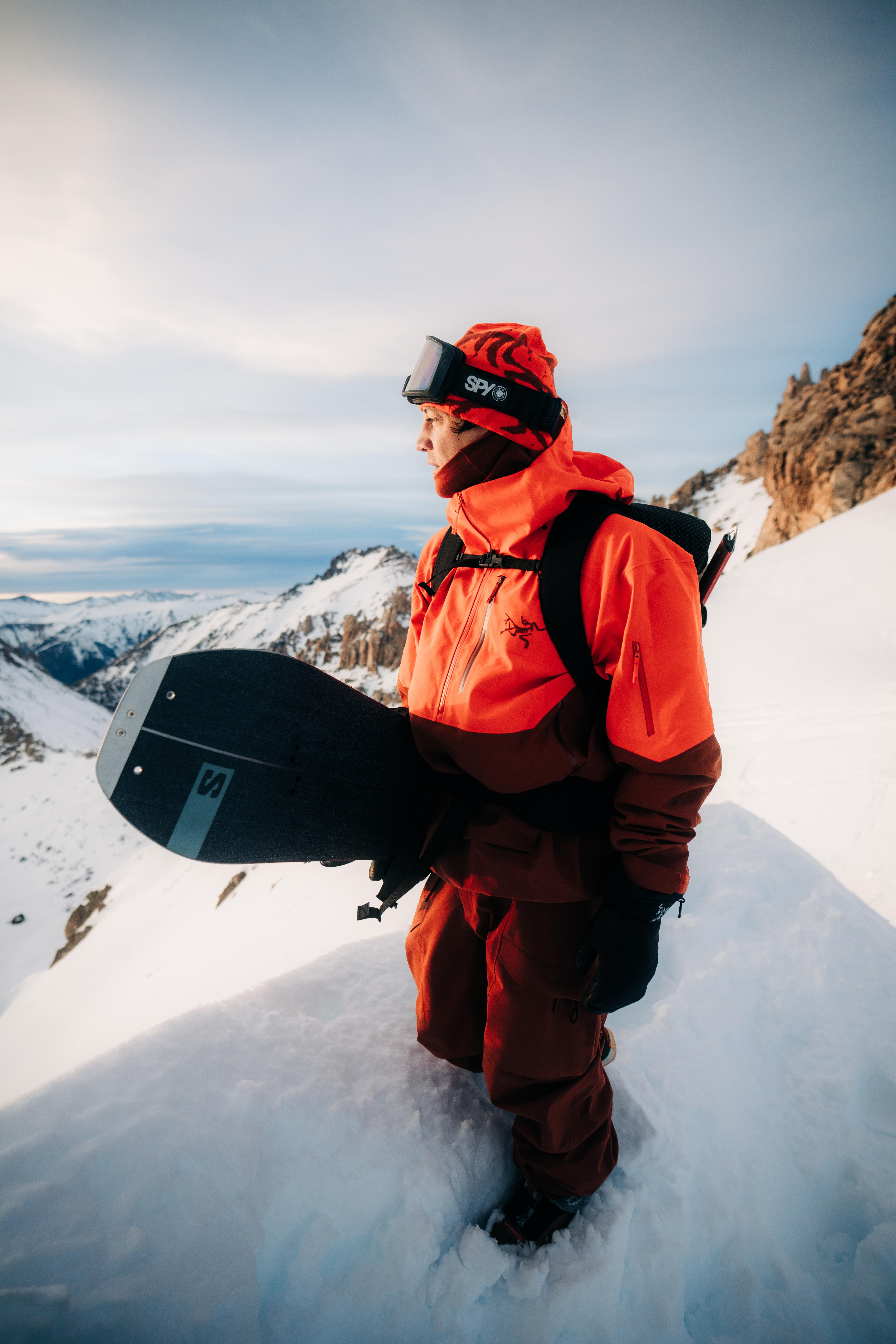 A snowboarder in orange winter gear stands on a snowy mountain ridge, holding a snowboard and looking out at the mountains.
