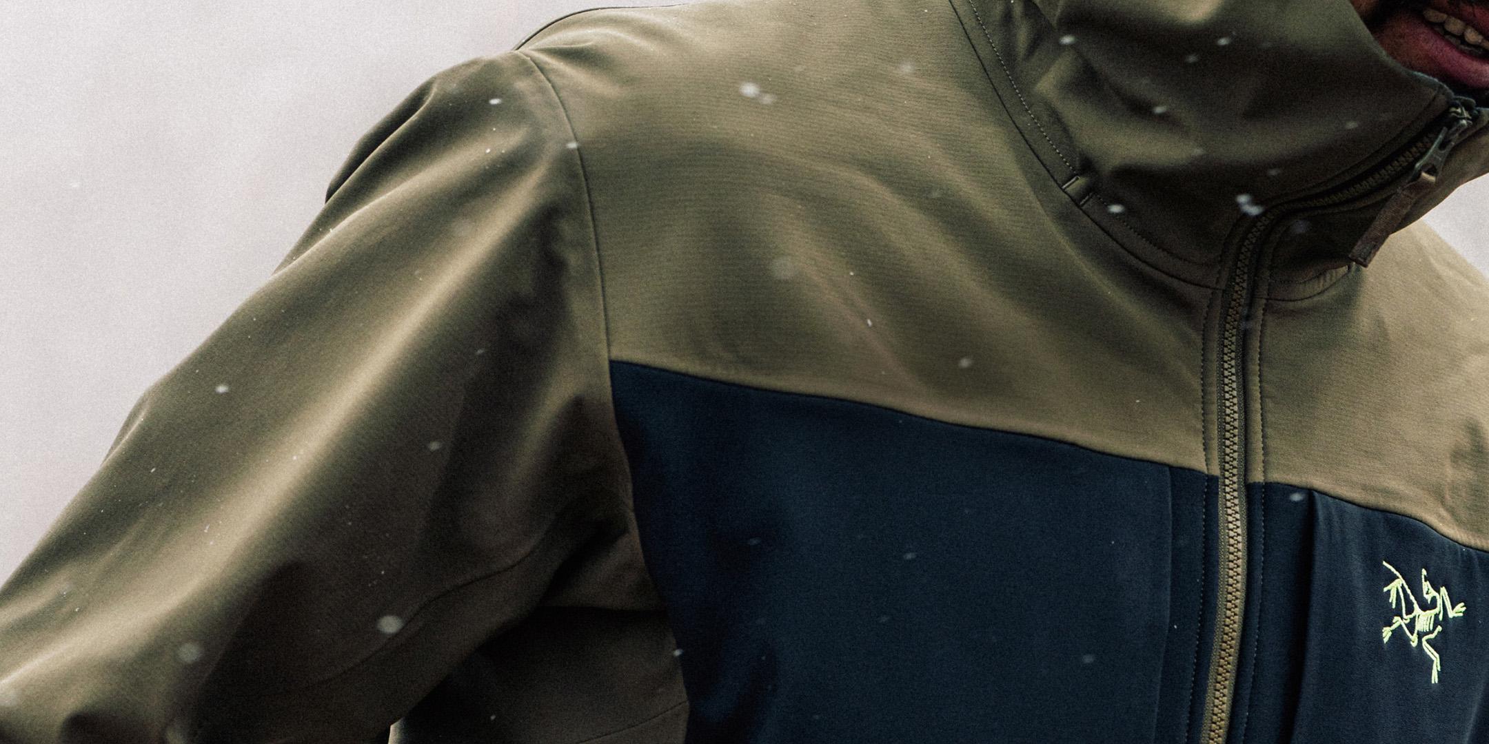 Person wearing an olive green and dark blue Arc'teryx jacket in falling snow.