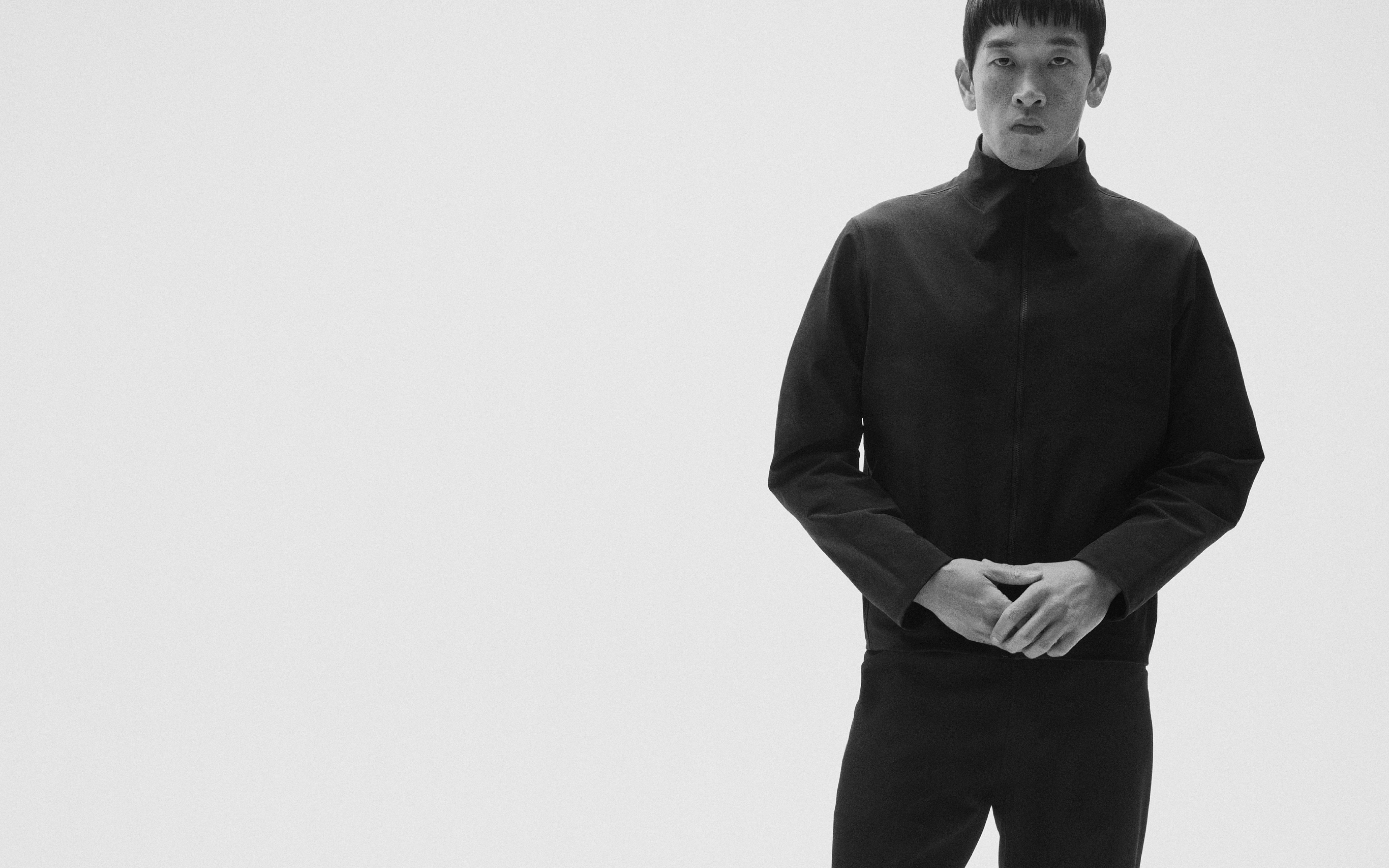 Black and white image of a man in a dark jacket and pants, hands clasped, against a white background.