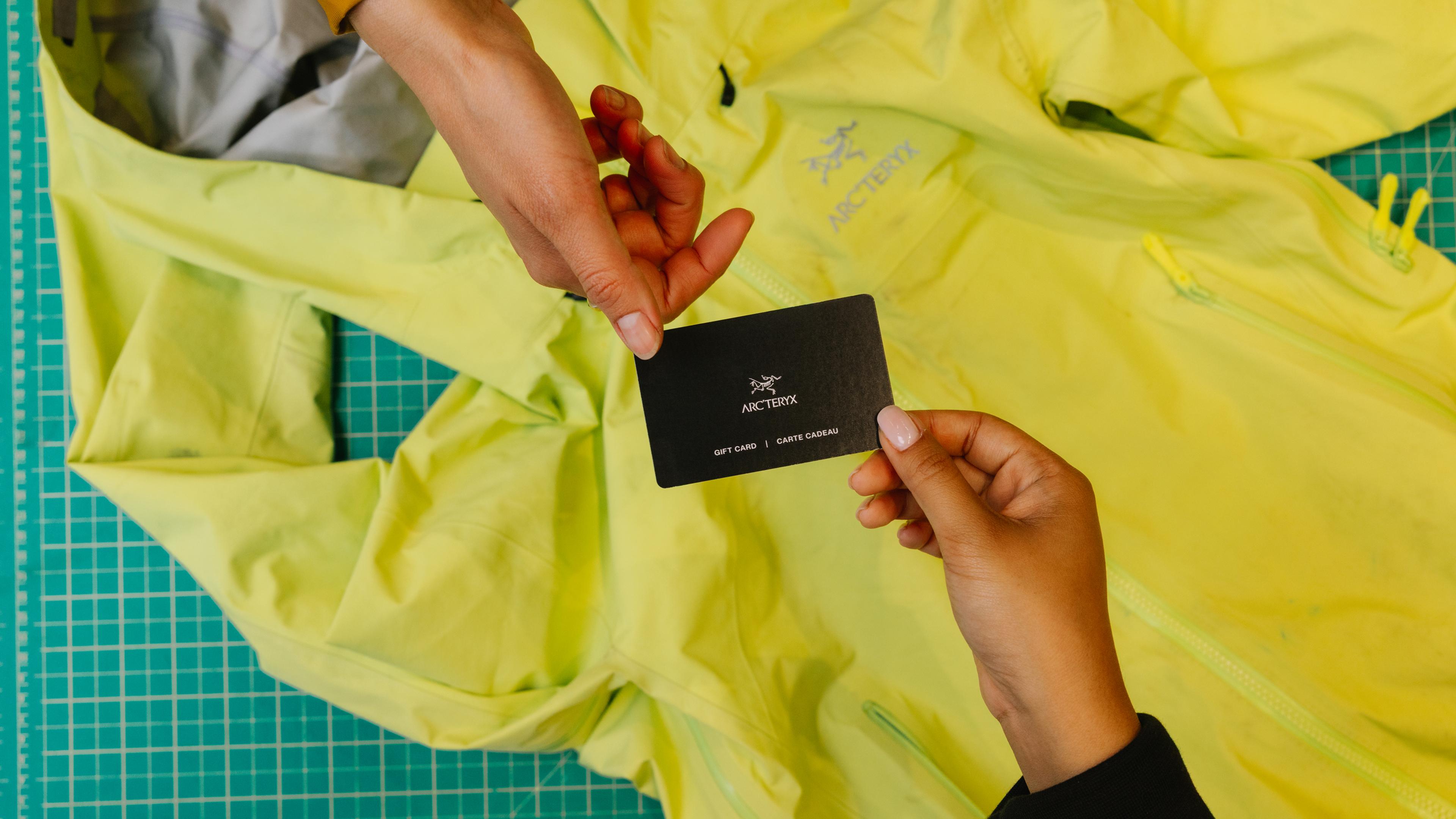 Two hands exchanging an Arc'teryx gift card over a yellow Arc'teryx jacket.