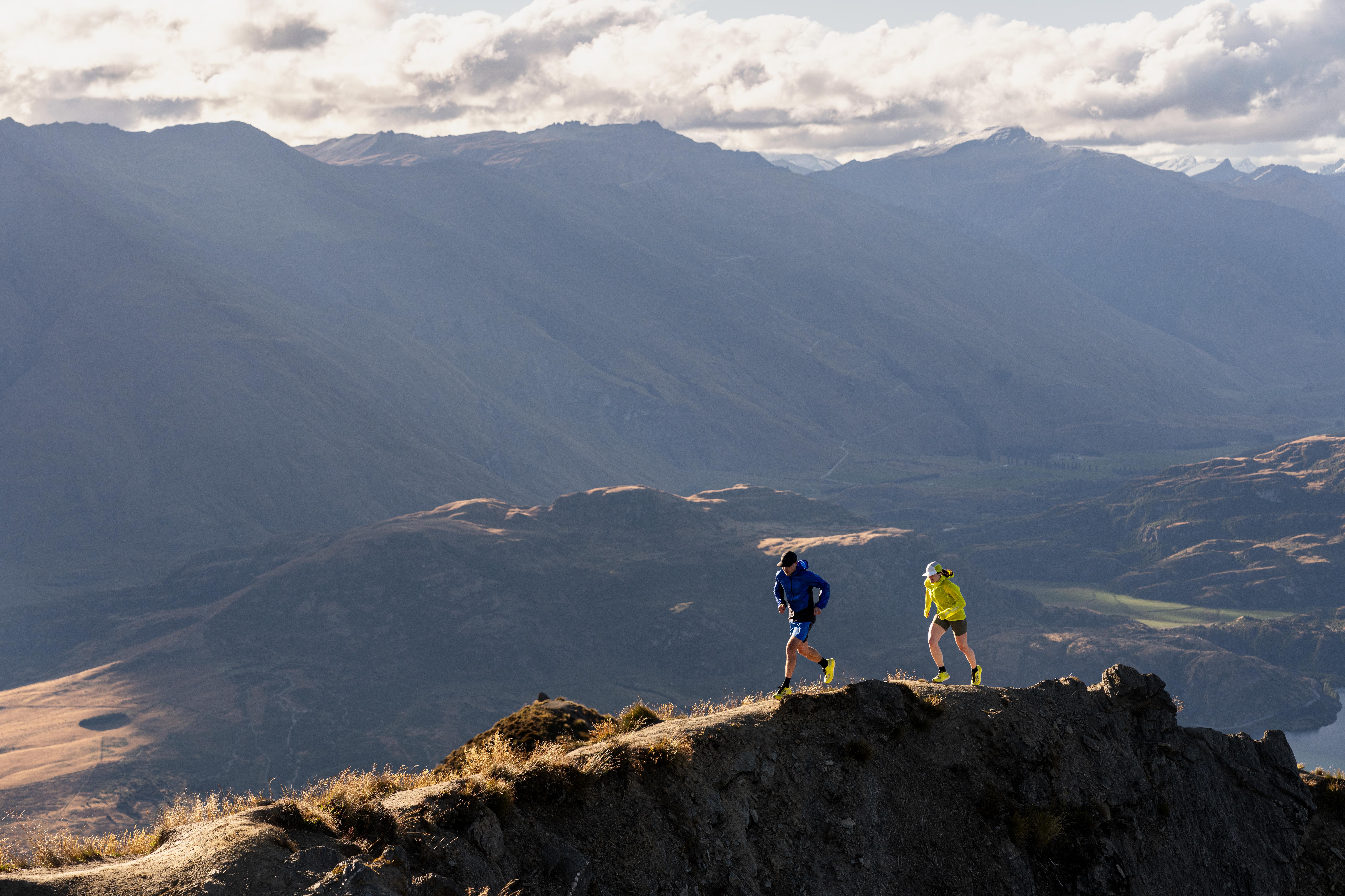 Two runners on a mountain ridge with a vast mountain range in the background.
