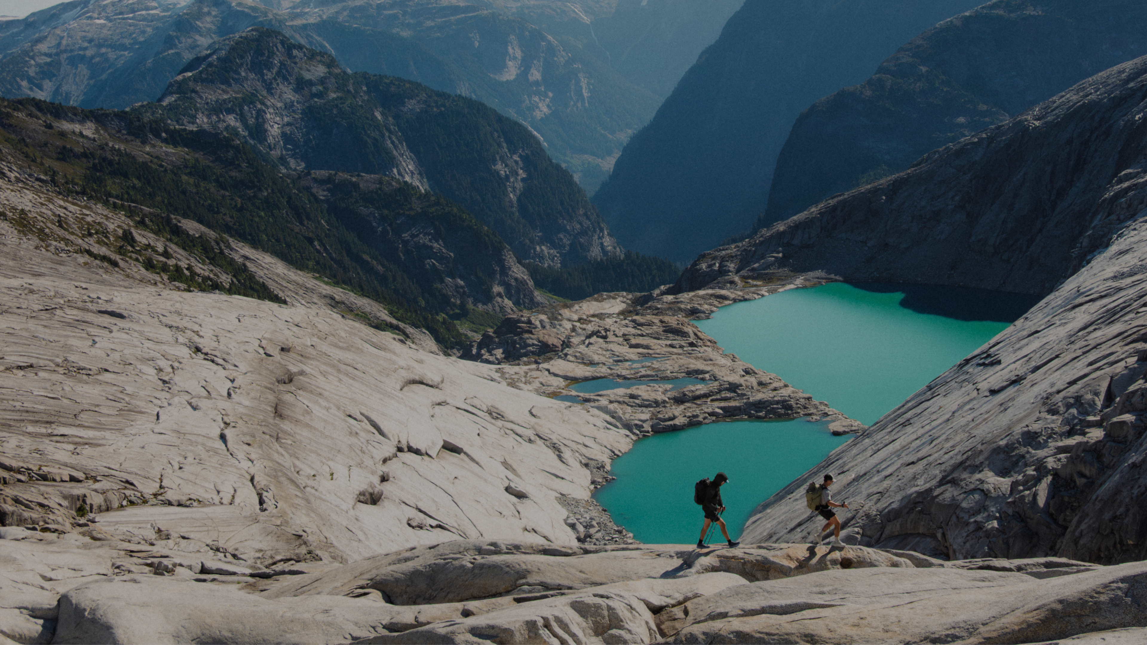 Two hikers walk on a rocky slope above turquoise lakes, with mountains in the background.