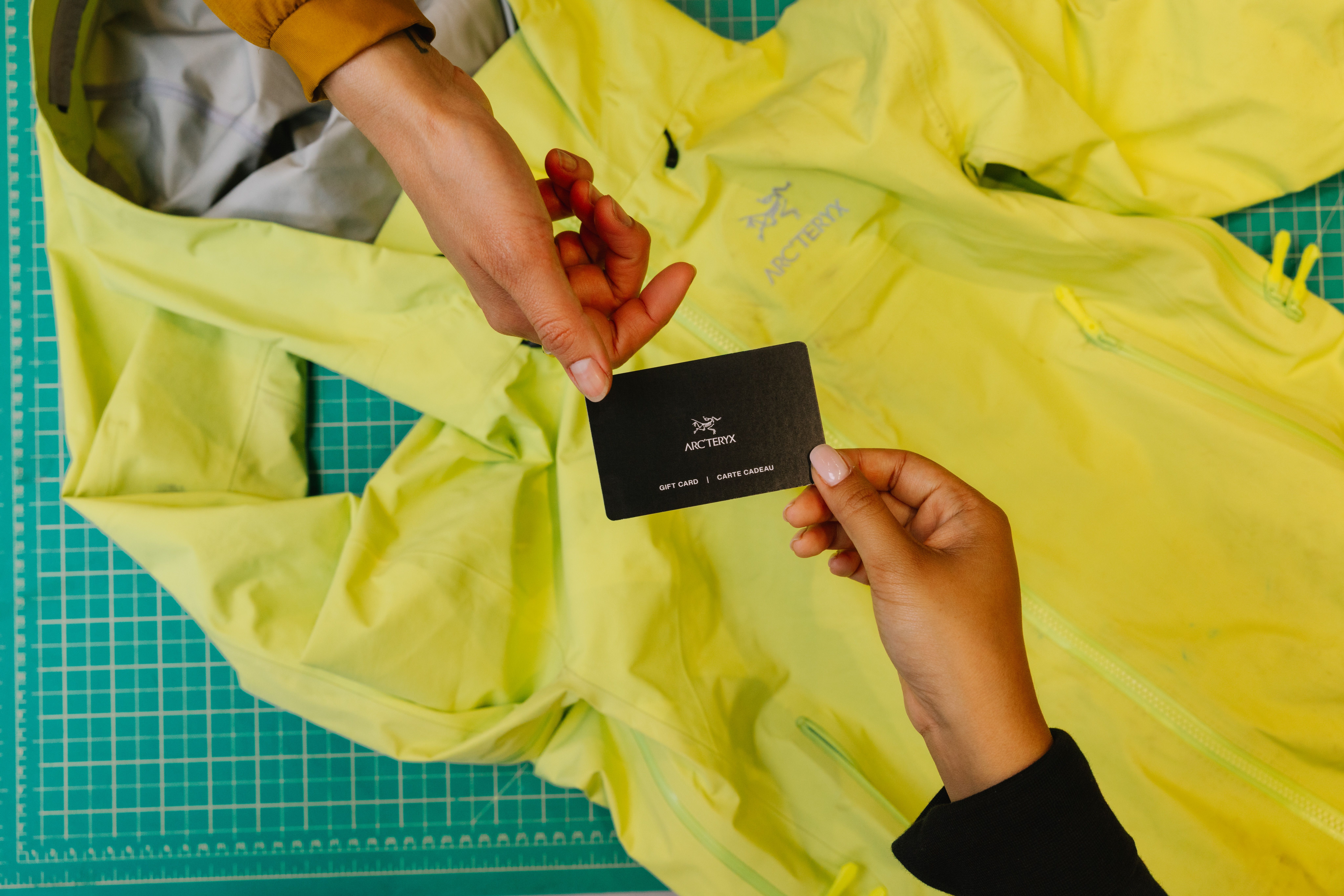 Hands exchanging an Arc'teryx gift card above a bright green jacket.