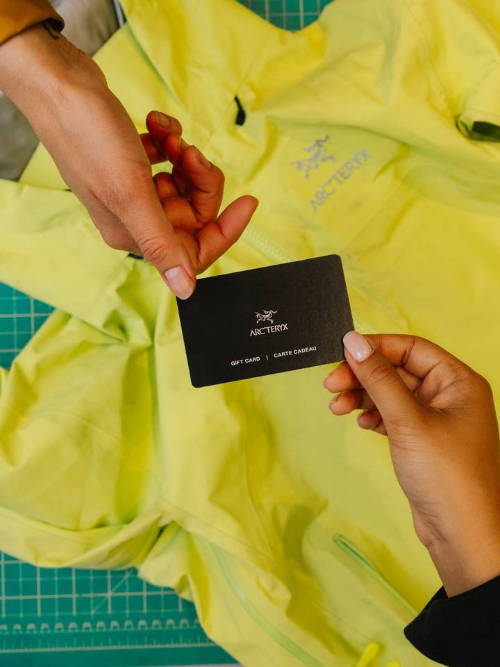 Hands exchanging an Arc'teryx gift card above a bright green jacket.