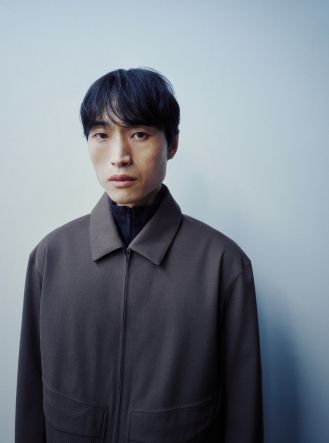 A man in a dark jacket and black turtleneck looks directly at the camera.