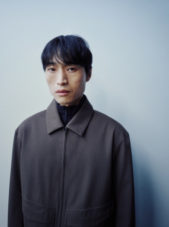 Portrait of an East Asian man in a dark grey collared jacket over a dark blue top, looking at the camera.