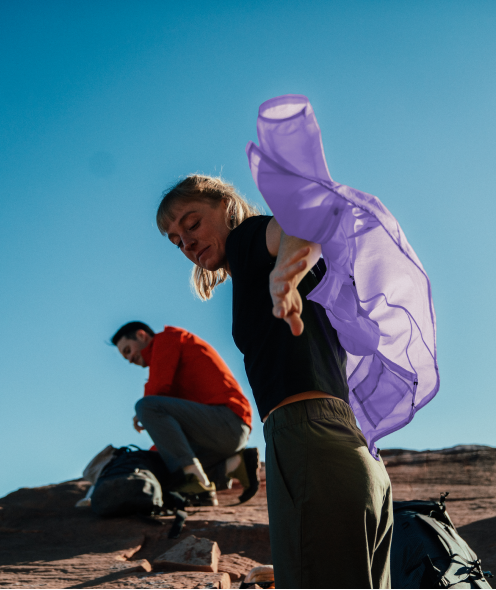 A woman outdoors with sheer purple fabric billowing in the wind, with a man crouching behind her on rocky ground under a blue sky.