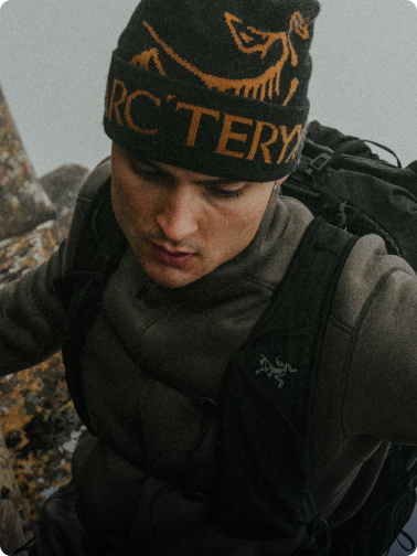 A man in an Arc'teryx beanie and backpack looks down intently while climbing.