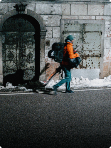 A person with skis and a backpack walks past an old building with rusty doors and snow.