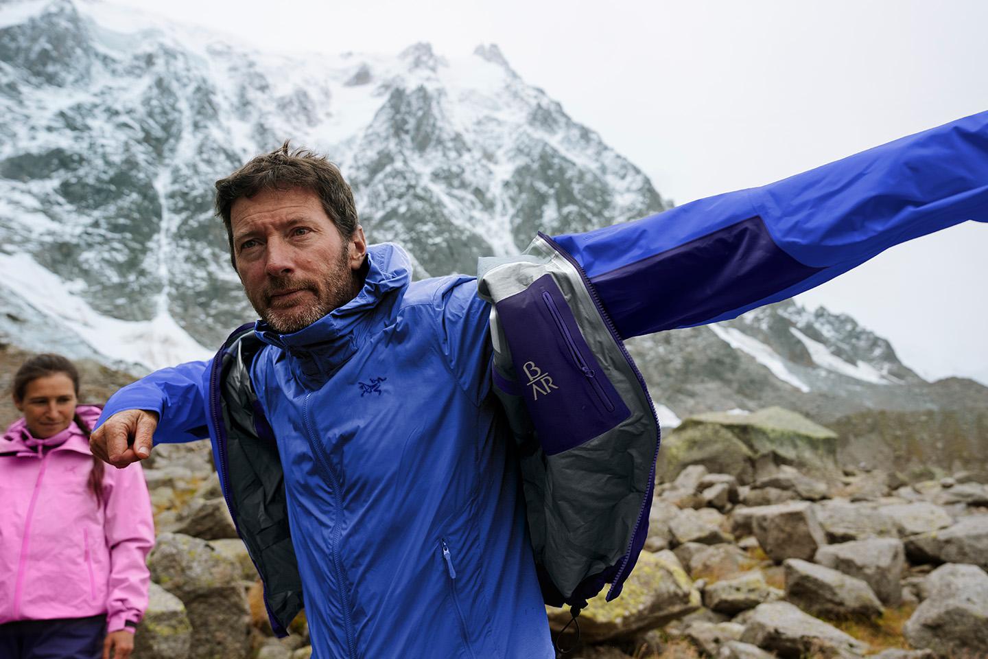 a man in a blue Beta AR jacket is standing in front of a mountain with his arms outstretched