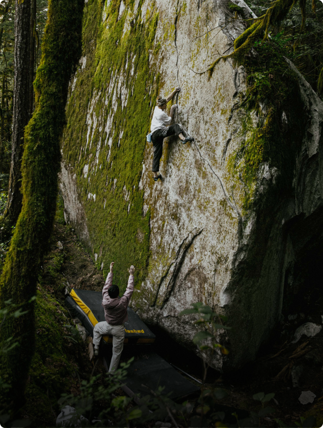 A person bouldering on a moss-covered rock with a spotter below.