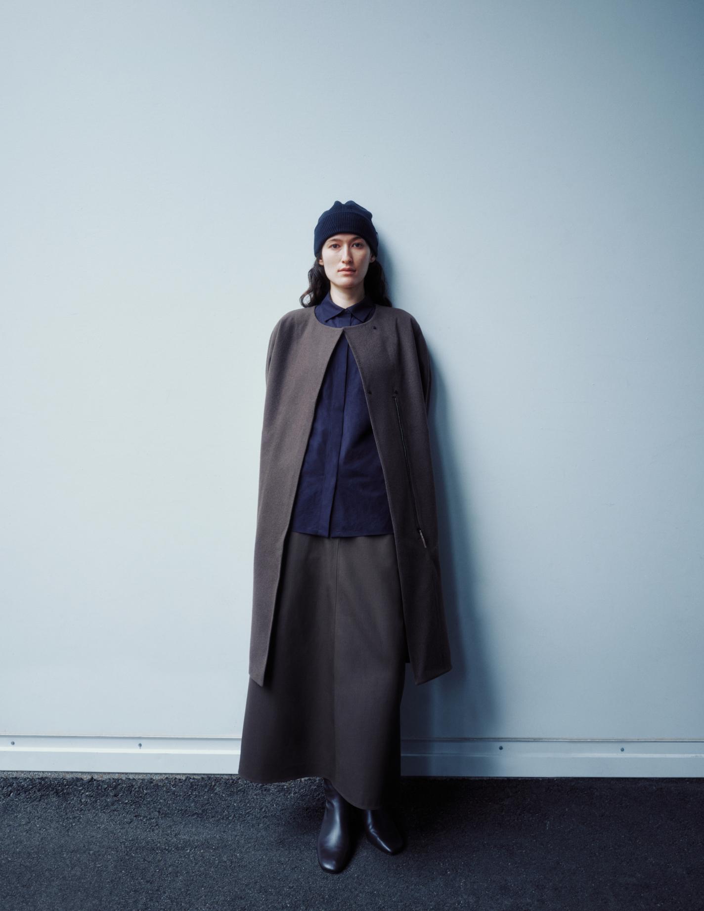 A woman in a dark beanie, dark blue shirt, long dark brown cape, and matching skirt stands against a light blue wall.