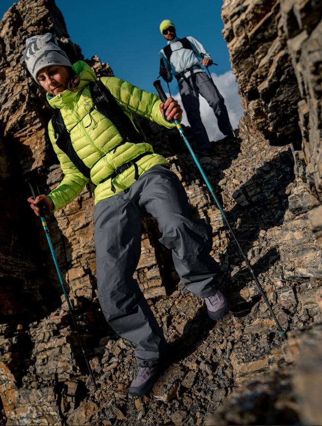 A woman in a neon green puffy jacket and a man behind her descend a rocky slope, both using trekking poles.