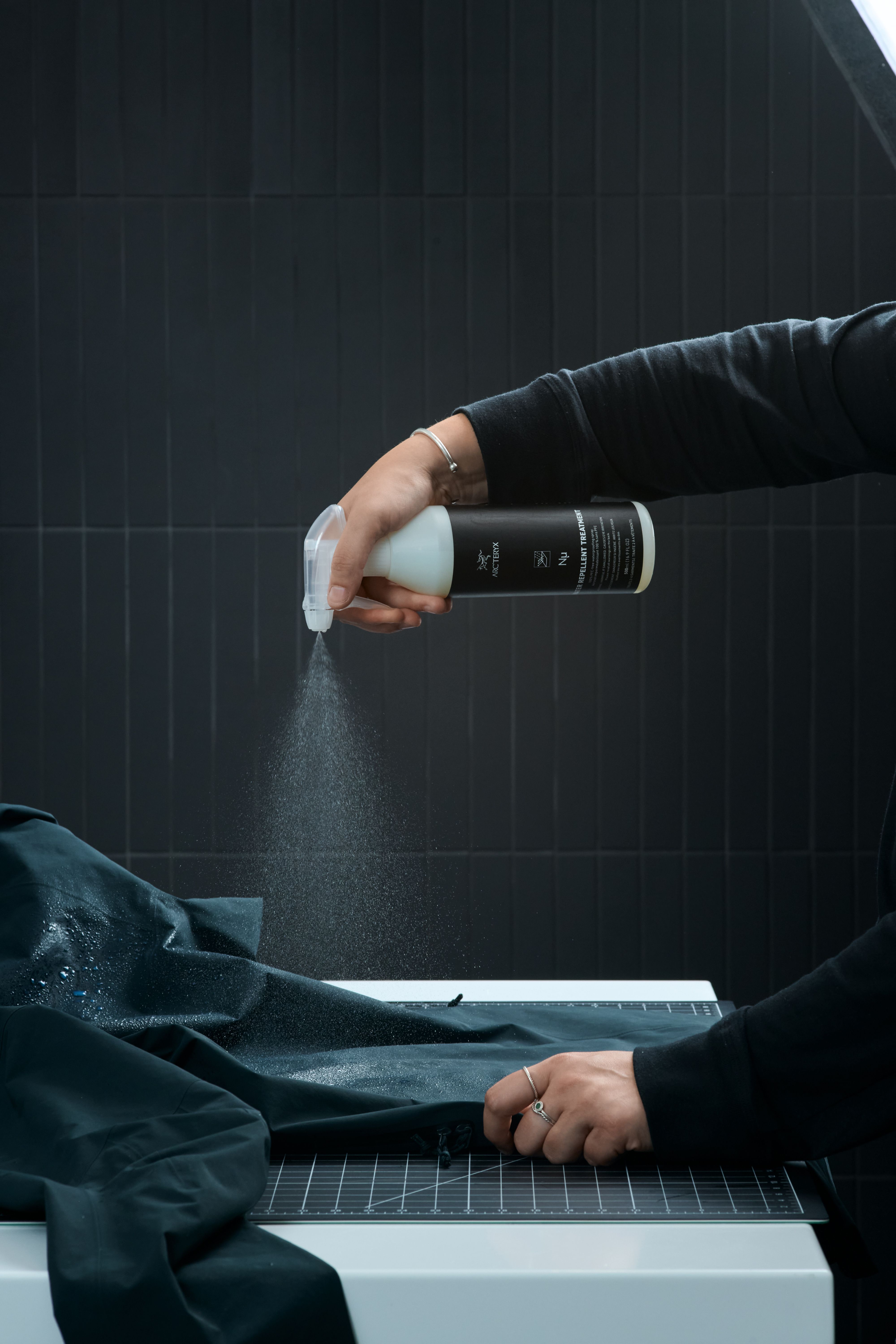 A person sprays Arc'teryx water repellent treatment from a bottle onto dark fabric, with visible mist and water droplets.