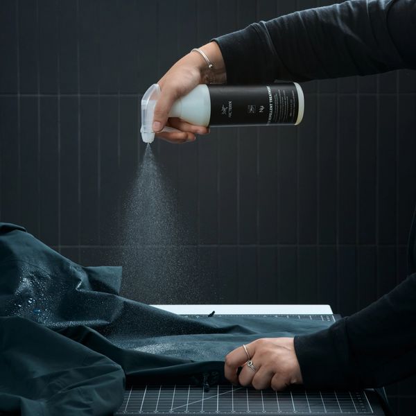 Hands spray an Arc'teryx water repellent treatment onto a dark technical jacket.