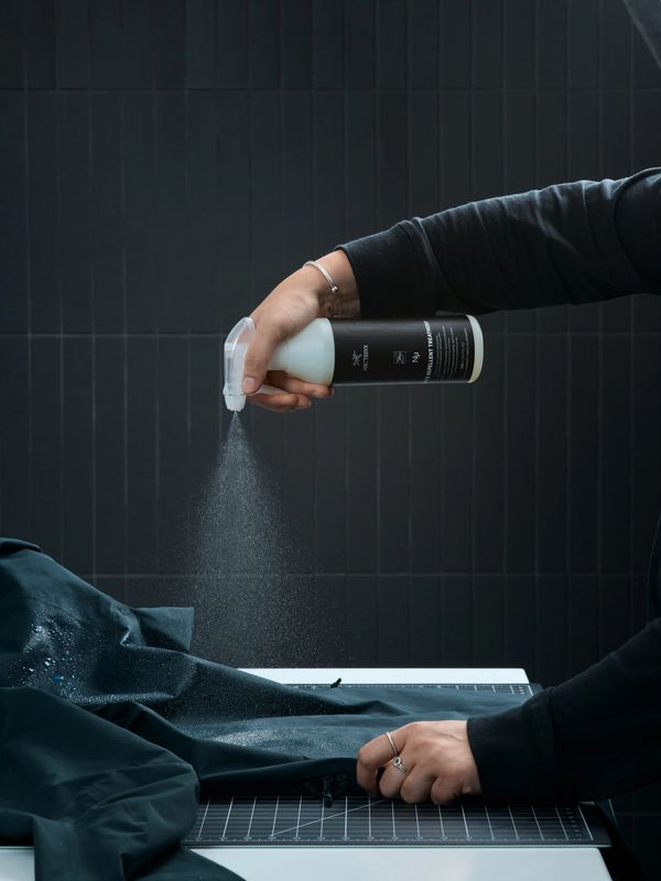 A person sprays Arc'teryx water repellent treatment from a bottle onto dark fabric, with visible mist and water droplets.