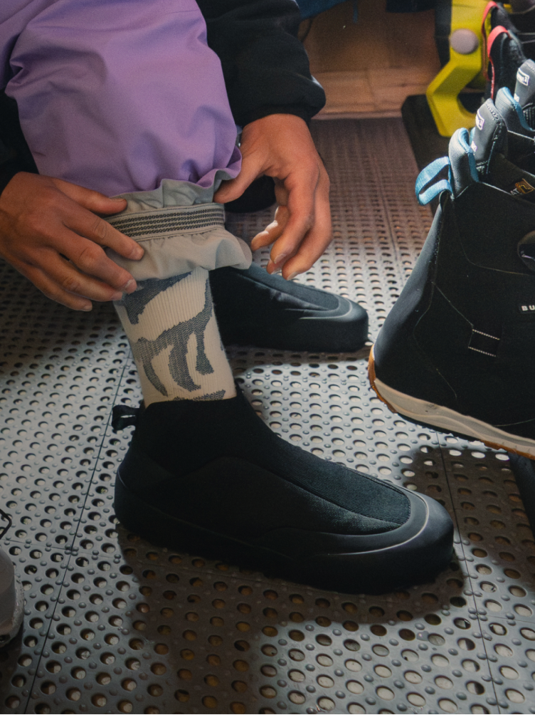 A person adjusts their pant cuff above a patterned sock and a black slip-on shoe on a perforated floor.
