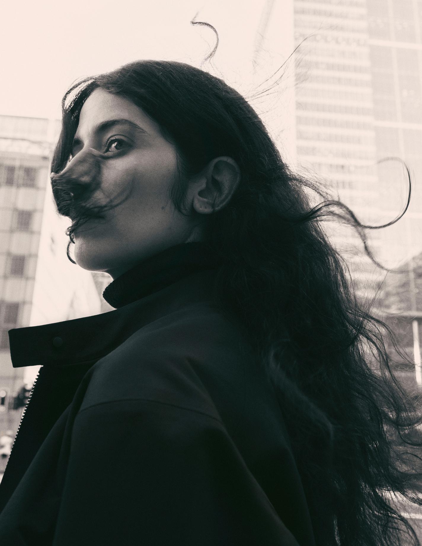A woman with windswept hair looks over her shoulder in a black and white city scene.