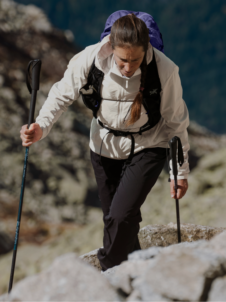 A person in a light jacket and dark pants, wearing a hydration vest, stepping on rocks.