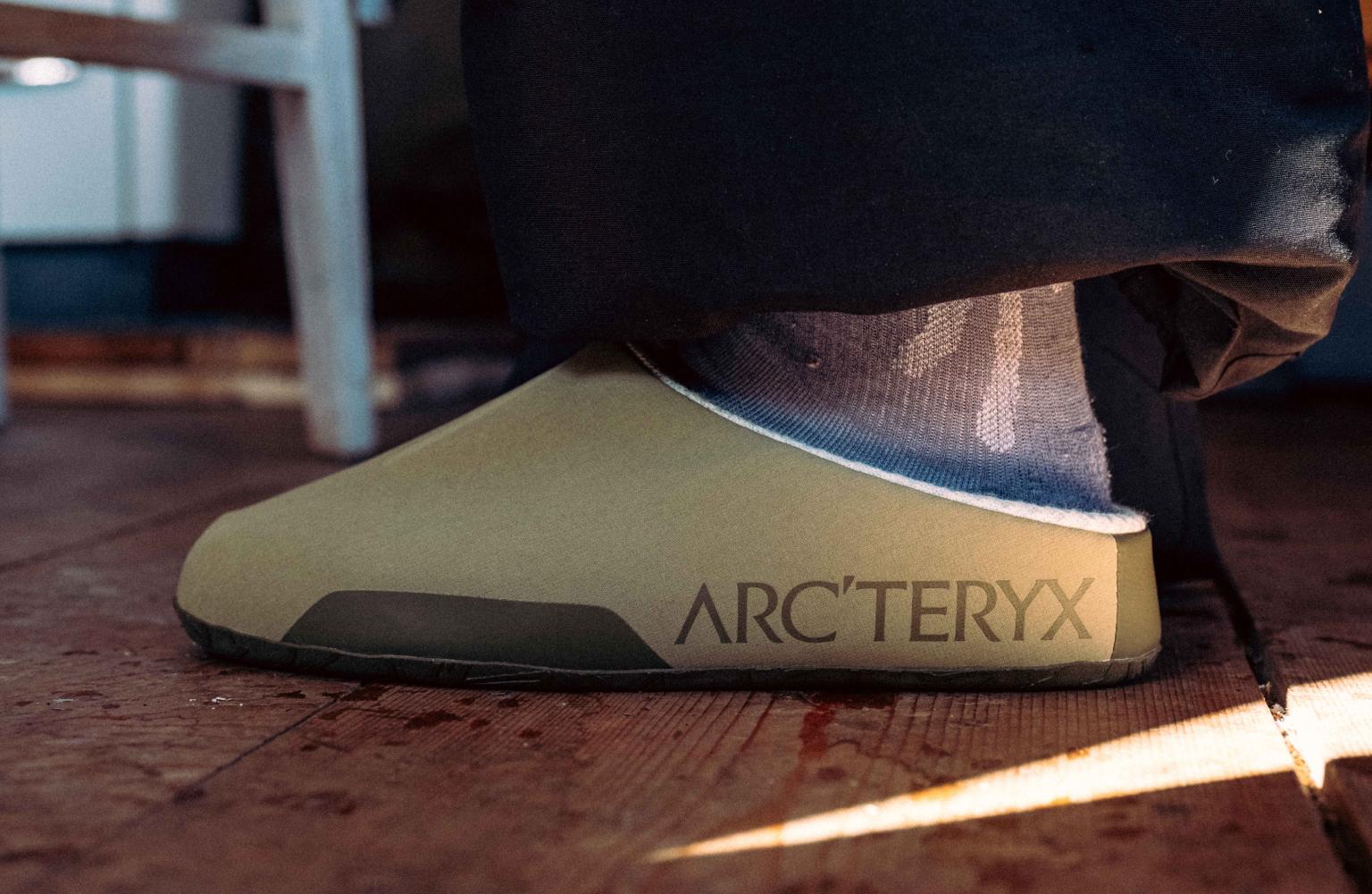 A foot in a tan Arc'teryx clog and sock on a wooden floor.