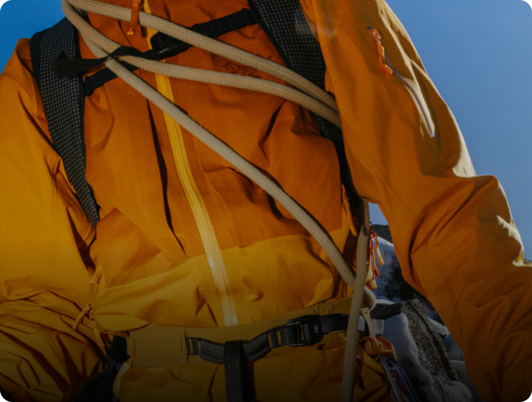 Climber in orange jacket with rope and backpack under a blue sky.