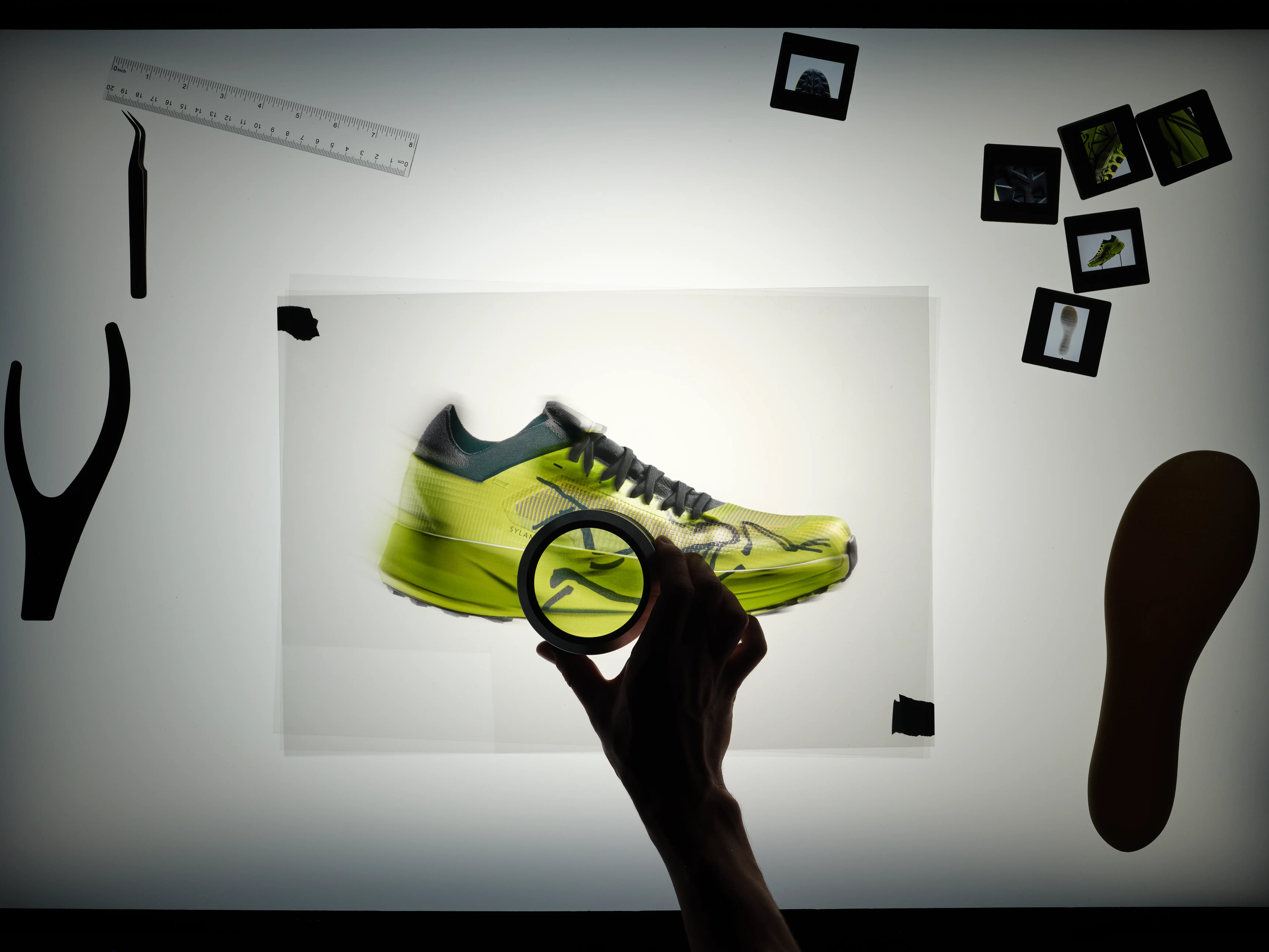 A hand with a magnifying glass examines a motion-blurred neon yellow running shoe photo on a lightbox surrounded by design tools and film slides.